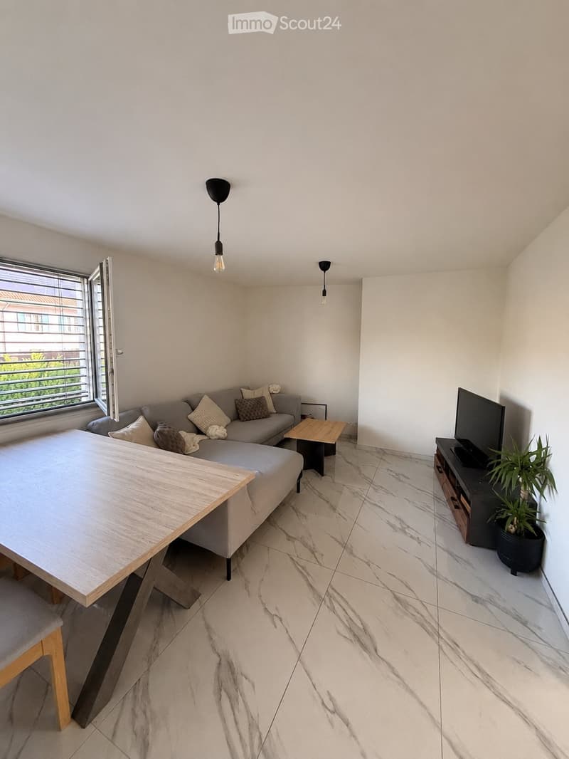 Modern living room with a grey sectional sofa, wooden coffee table, flat-screen TV on a stand, and a potted plant near the window. The room features marble flooring and large windows with blinds.