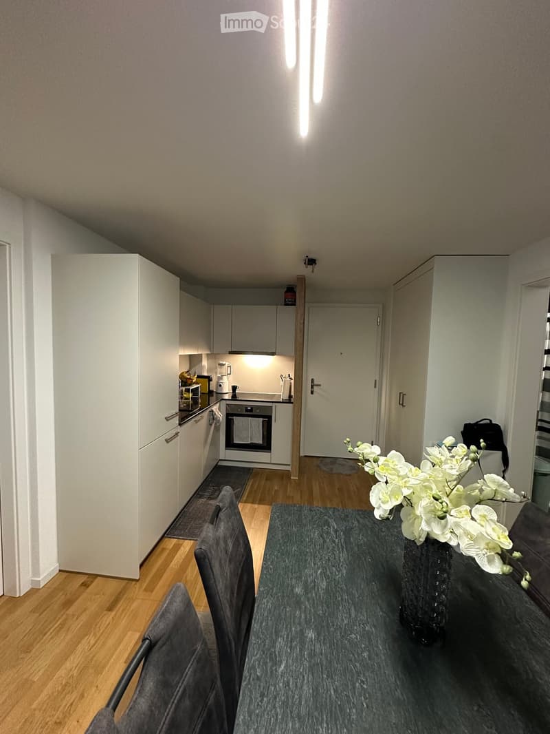 A modern, open-plan living and dining area with a grey dining table, chairs, a sofa, and a vase with white flowers. The kitchen area features white cabinets, an oven, and a refrigerator.