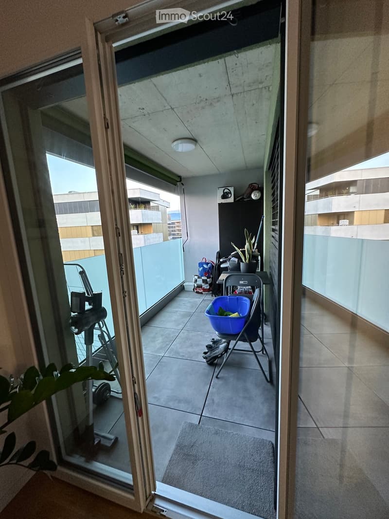 Balcony with tiled floor, glass railings, and a view of the building exterior. Includes a folding chair, a blue laundry basket, a small table, and a potted plant.