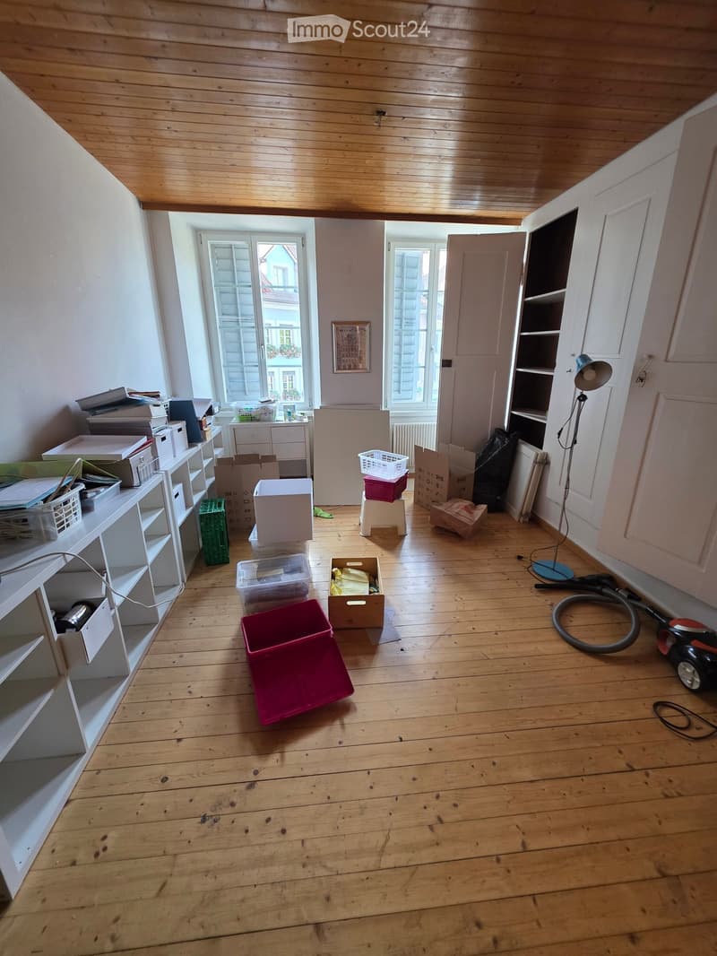Empty room with wooden flooring, white walls, and large windows. The space contains storage boxes, a vacuum cleaner, and a floor lamp, but no furniture.