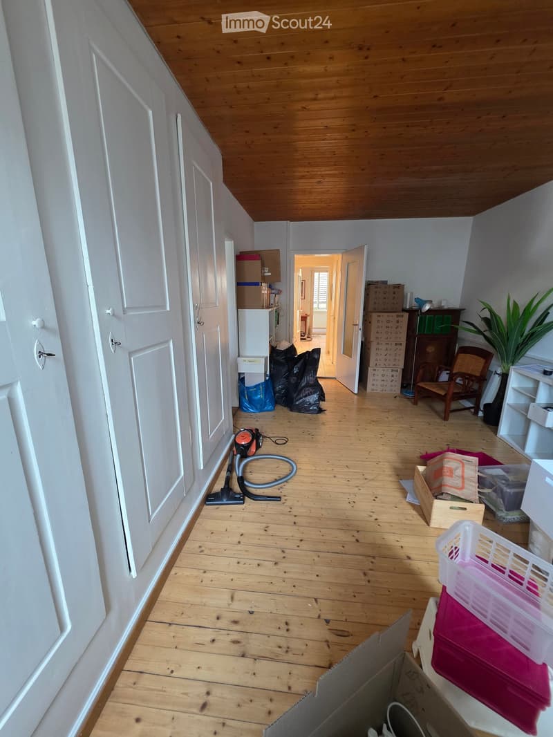 Empty wooden-floored room with white walls, several doors, and scattered items including a vacuum cleaner, boxes, and a chair.