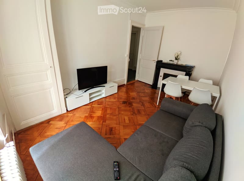 A modern living room with a grey sofa, a flat-screen TV on a white stand, a dining table with four white chairs, and a small black fireplace with a vase on top.