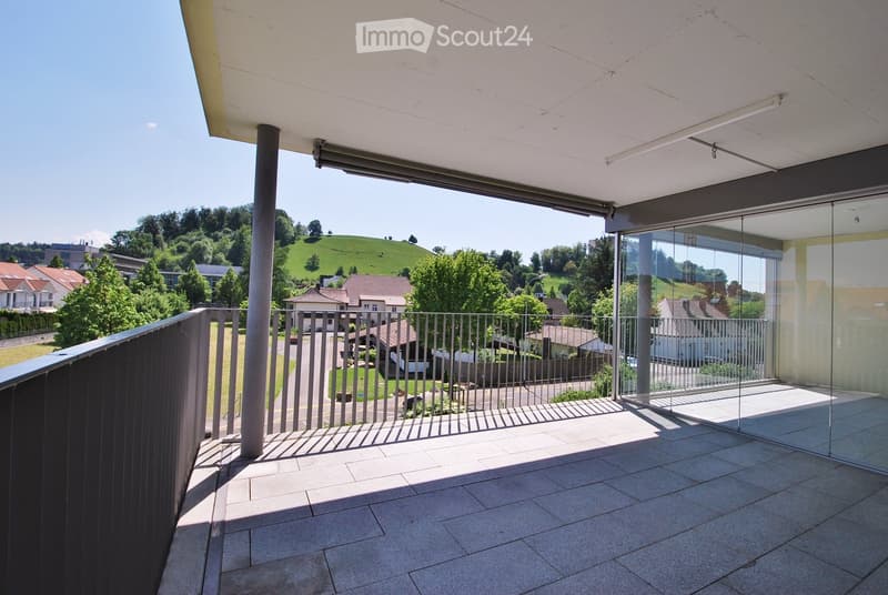 Modern balcony with tiled floor, glass railing, and metal balustrade, overlooking a suburban neighborhood with houses and greenery.