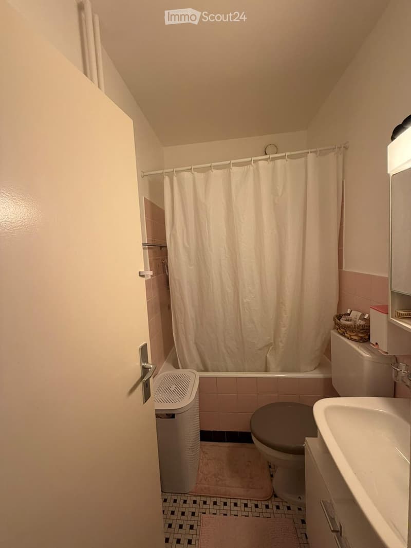 Bathroom with a toilet, sink, bathtub, shower curtain, and a small trash can. The room features tiled walls and a patterned floor.