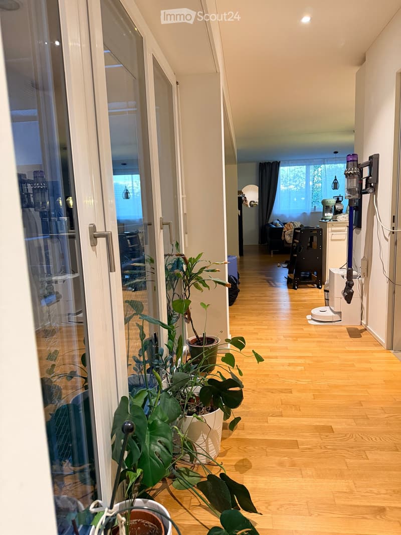 A well-lit hallway with wooden flooring, large potted plants near the entrance, and a view into an adjacent living area with a sofa and a window.