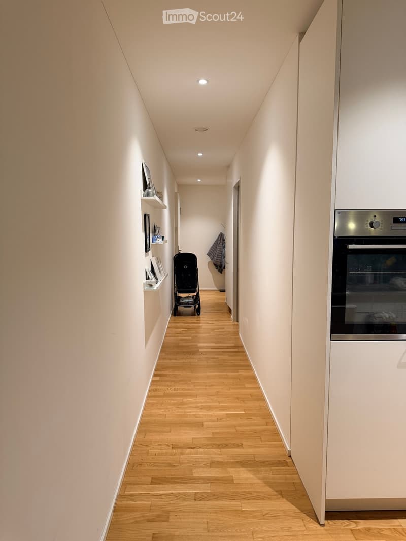 A narrow hallway with light wooden flooring, white walls, and built-in shelves on one side. A stroller is positioned in the center, and a modern oven is visible on the right side.