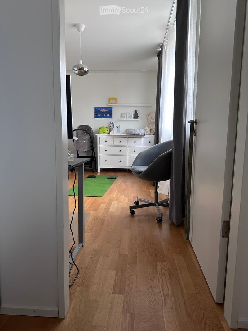 A well-lit room with wooden flooring, a grey chair, a small table, a drawer unit, a baby stroller, and a shelf with toys and decorations.