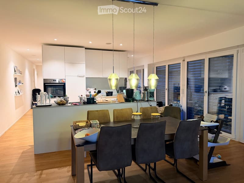 Modern kitchen with white cabinets, a dining area, and pendant lights. The kitchen features a countertop with various appliances and utensils, and a dining table with chairs and a baby chair.