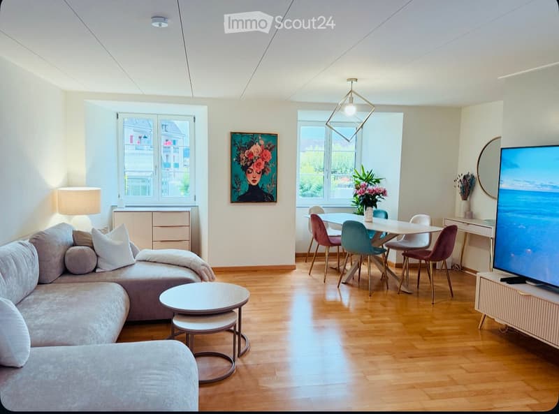 A modern, open-concept living and dining area with a grey sectional sofa, a small coffee table, a dining table with colorful chairs, a television on a stand, and a painting on the wall. The space features wooden flooring and large windows with a view of t