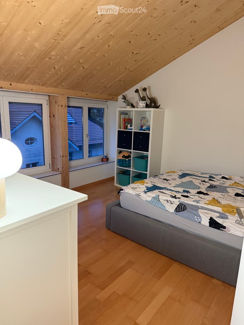 Cozy attic bedroom with wooden slanted ceiling, hardwood floor, large windows, bed with colorful bedding, shelf with books and decorative items, and a modern lamp on the left side.