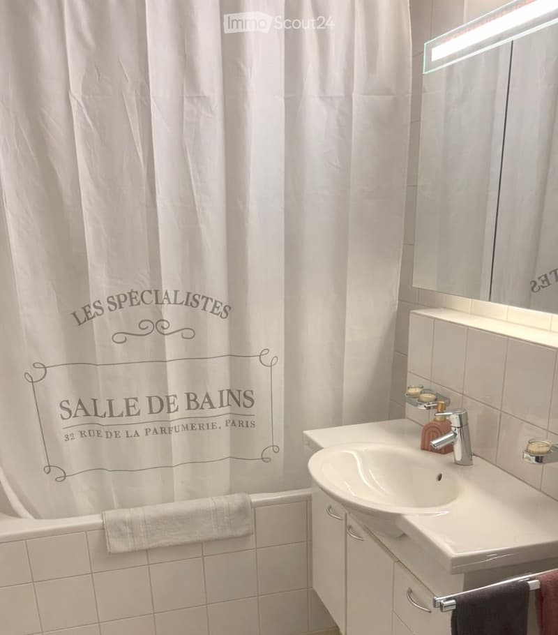 Modern bathroom with a white bathtub, shower curtain, sink, and mirror. The space features white tiles and a sleek design.