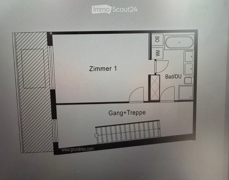 Floor plan showing a room labeled "Zimmer 1", a bathroom/toilet area, a hallway/stairs area, and a small storage or closet space.