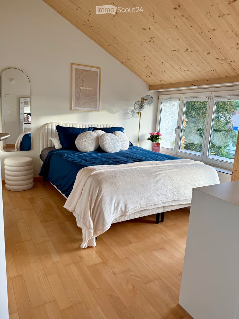 Cozy attic bedroom with wooden floors, slanted wooden ceiling, large windows, a neatly made bed with dark blue bedding, decorative pillows, a round mirror, a small stool, and a modern lamp.