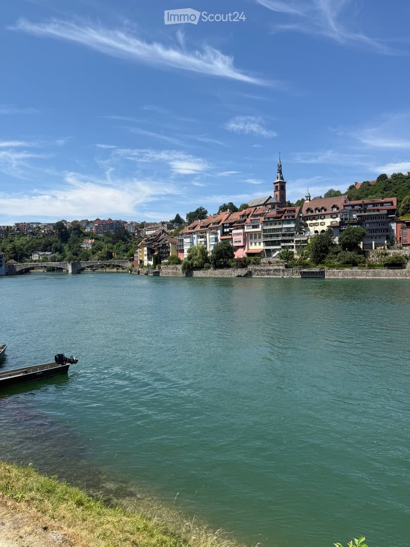 A scenic view of a river with a small boat on the left, a bridge in the background, and a town with colorful buildings and a church tower on the right.