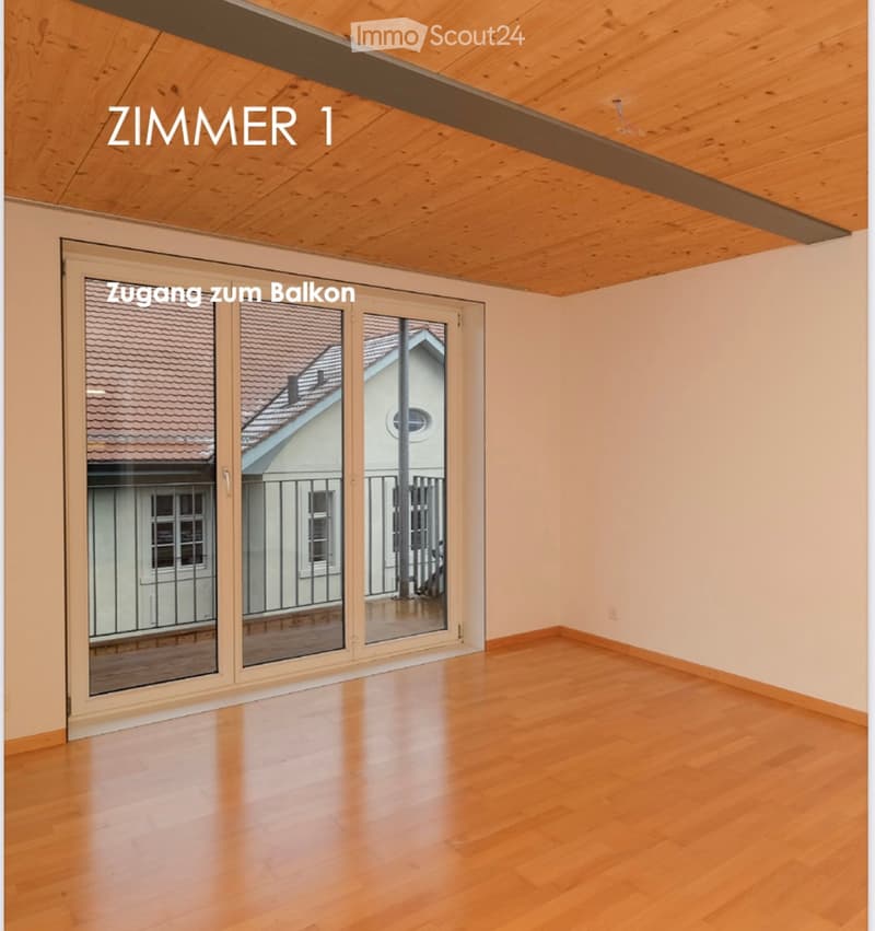 Empty room with wooden flooring, large sliding glass door leading to a balcony, and exposed wooden ceiling beams.