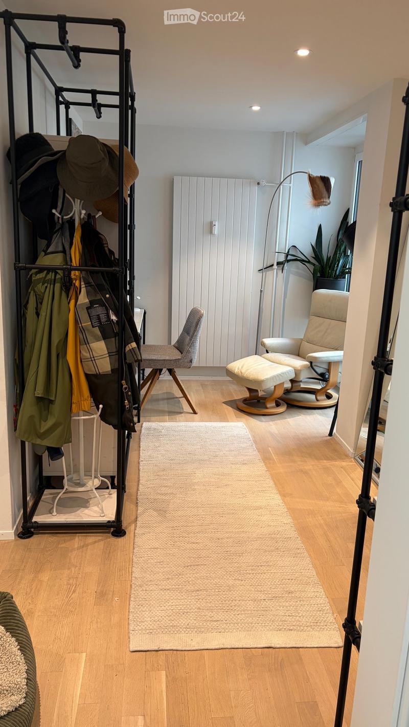 A modern hallway with wooden flooring, a cream-colored rug, a coat rack with jackets and hats, a small table, a chair, a stool, and a floor lamp.