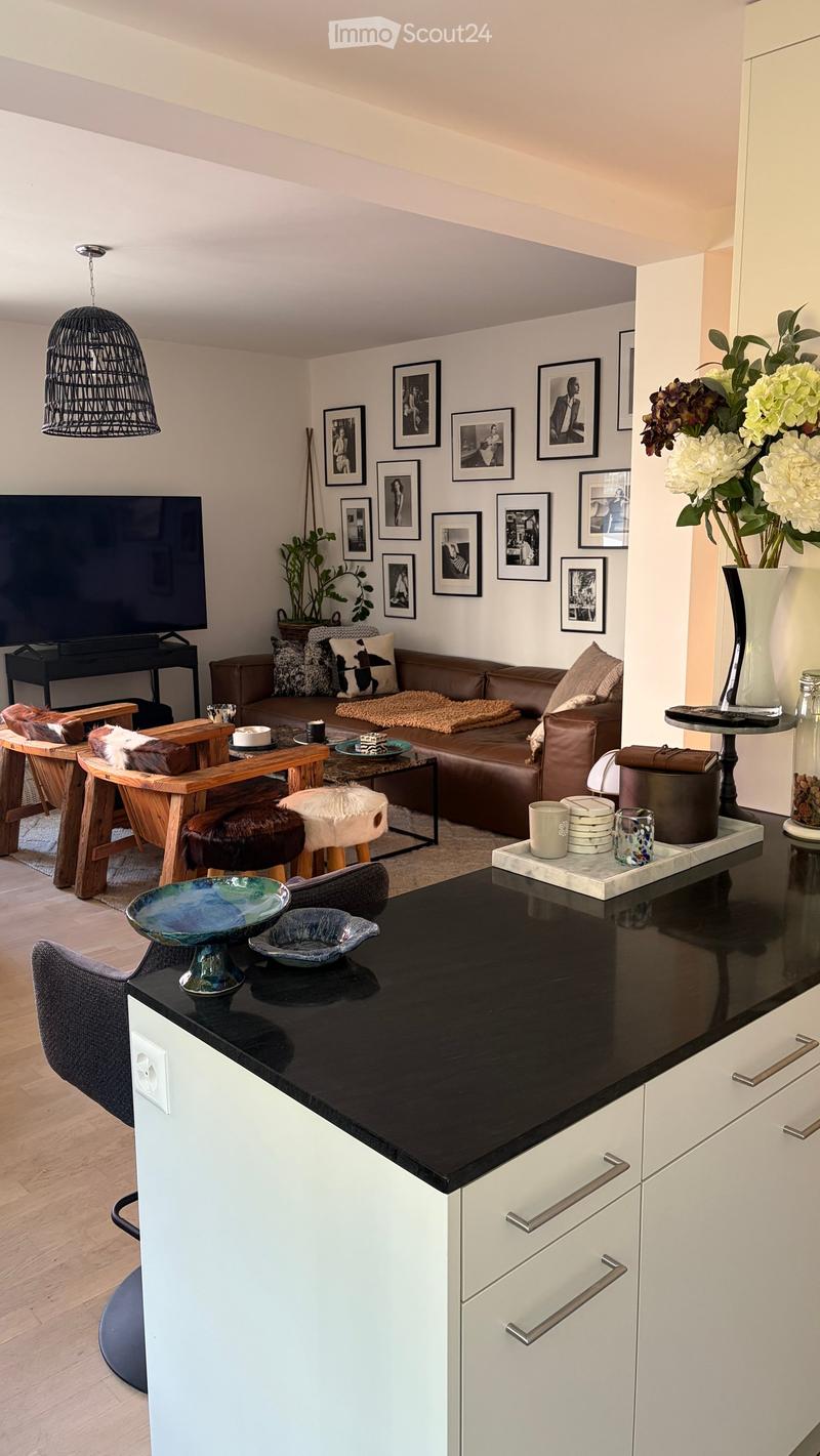 Modern living room with a brown leather sofa, wooden coffee table, decorative bowls, and a pendant light. The space includes a kitchen area with white cabinets and a black countertop.