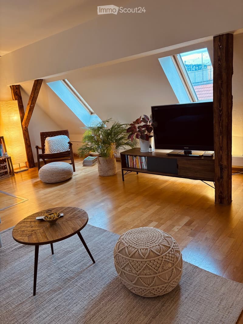 Modern living room with wooden floors, large windows, a television on a stand, a round coffee table, two ottomans, a chair, and a floor lamp.