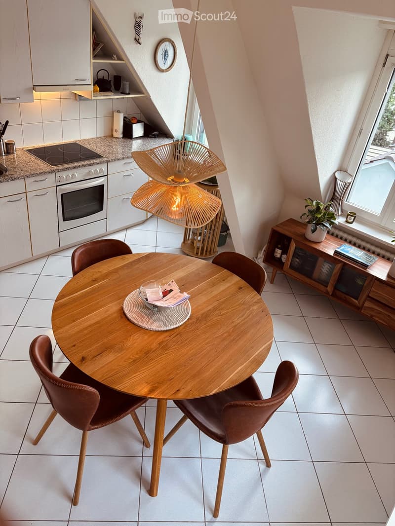 A modern, well-lit kitchen and dining area with a round wooden table, four brown leather chairs, a wicker hanging lamp, and a kitchen area with white cabinets, a stove, and a countertop.