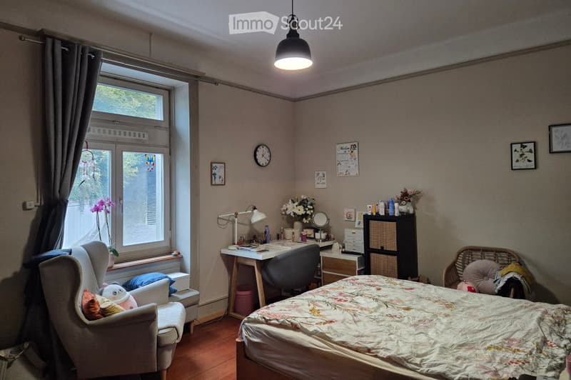 Cozy bedroom with a bed, desk, chair, and decorative items. Features a large window with curtains, a hanging pendant light, and a wicker chair with pillows.