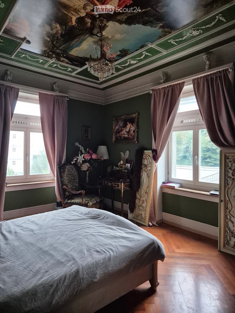 Elegant bedroom with a large bed, ornate chair, antique table, chandelier, and decorative painting on the wall. Green walls, wooden floor, and large windows with curtains.