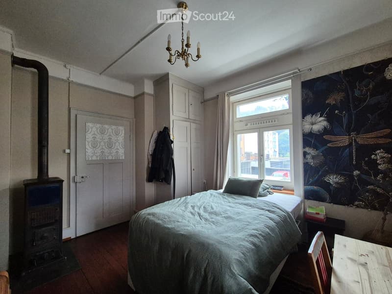 Cozy bedroom with a bed, wooden table, chair, cabinet, chandelier, and a fireplace. The room features a large window with a view and a decorative wall mural.