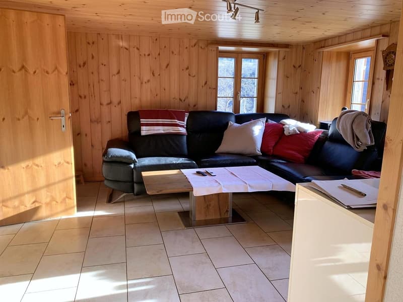 Cozy living room with wooden walls and ceiling, featuring a black leather sofa with colorful cushions, a wooden coffee table with a white table runner, and a small side table with papers and a pen.