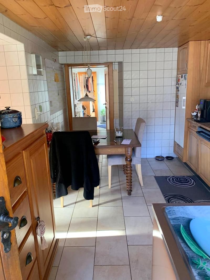 A well-lit kitchen and dining area with tiled walls and floors, wooden cabinets, a dining table with chairs, and various kitchen appliances.