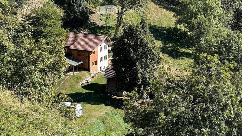 A two-story house with a red-tiled roof and white walls, surrounded by lush greenery and trees. The house features a covered patio area and is situated on a raised ground floor with a small garden and additional structures nearby.