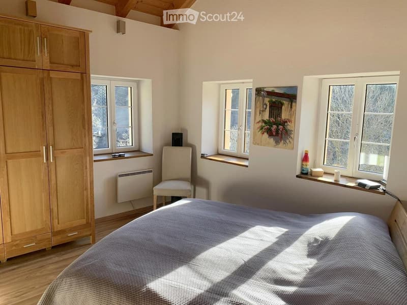 Bright bedroom with wooden floor, large windows, bed with blue bedding, wooden wardrobe, chair, and a painting on the wall.