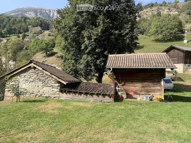 A rural outdoor area with a wooden cabin, stone structure, and a small table with chairs and a flower pot.