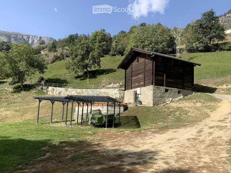 A wooden cabin sits on a stone base with a covered outdoor seating area featuring a table and chairs, surrounded by a grassy area and trees with a mountain view in the background.