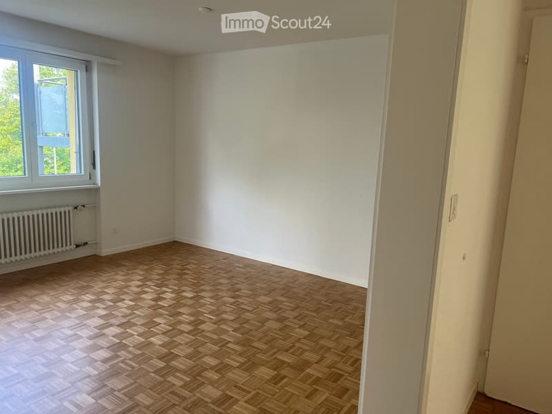 Empty room with wooden herringbone flooring, white walls, and a window with a radiator below it.