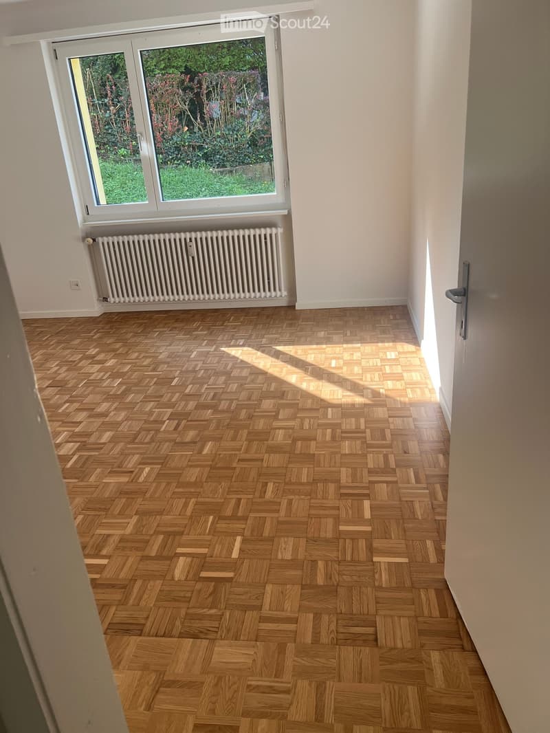 Empty room with herringbone wooden flooring, white walls, large window with view of garden, radiator, and door.