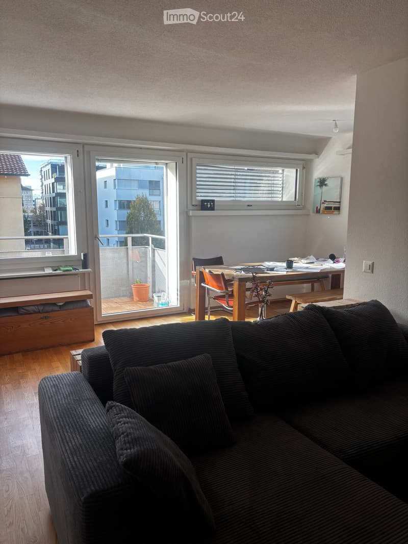 A modern living room with a dark grey sofa, a wooden dining table with chairs, a vase with flowers, and a balcony view of the city.