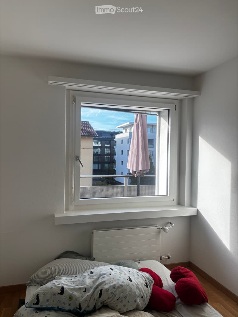 Bedroom with a bed covered in a patterned duvet and red pillows, a radiator below a large window with a view of modern buildings, and wooden flooring.