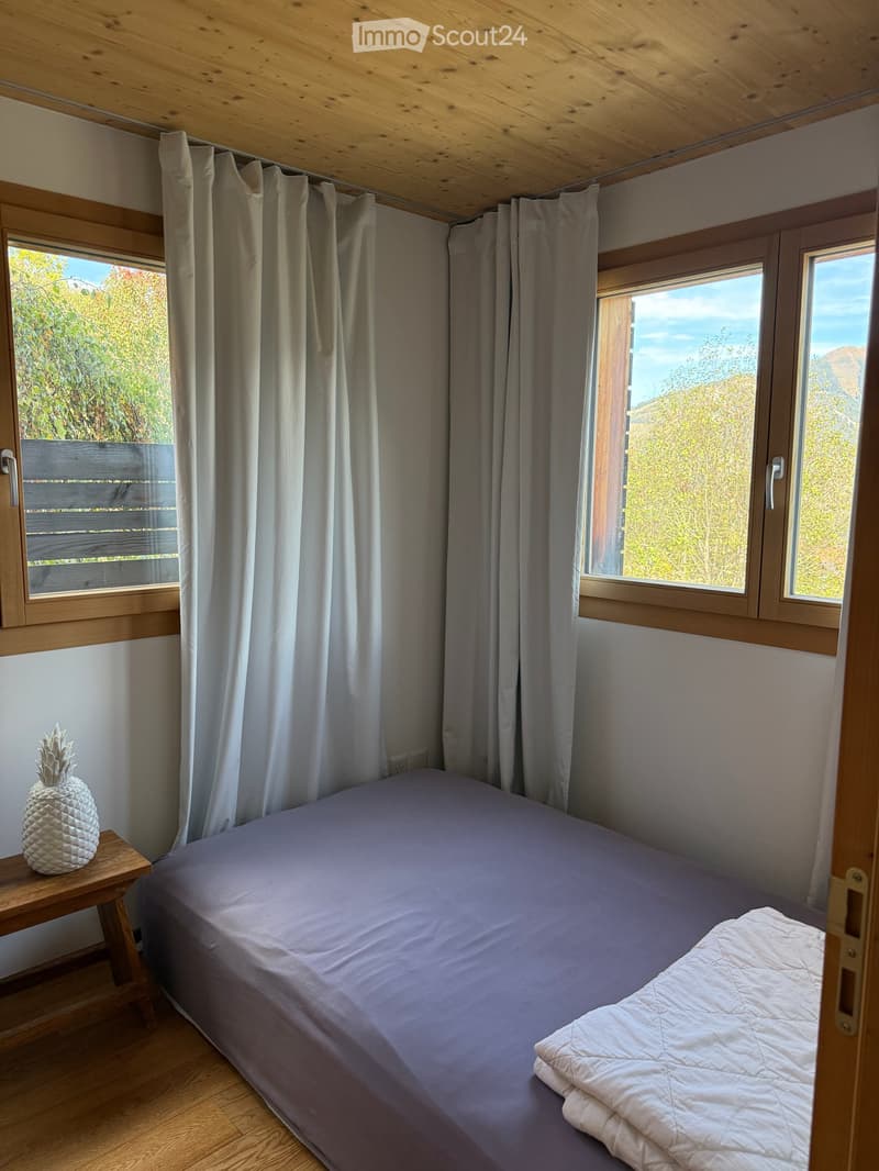 Cozy bedroom with a single bed, wooden nightstand, and a decorative pineapple on top. Large windows offer scenic views, and the room features light wooden ceilings and walls.