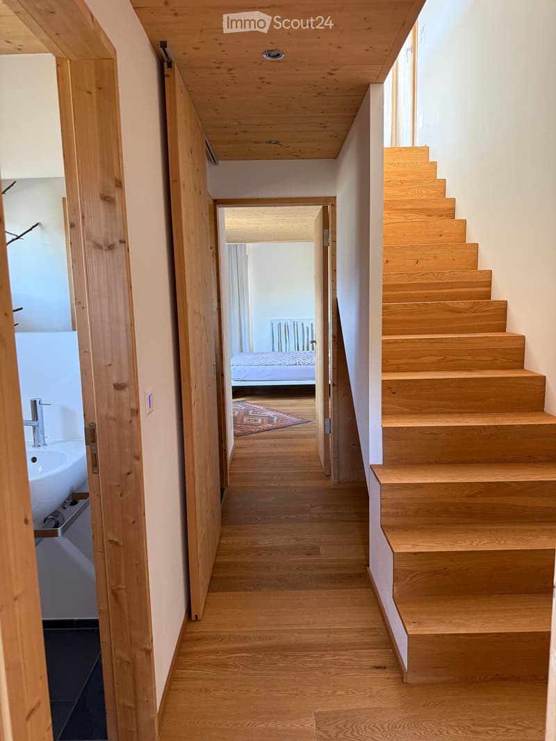 Wooden staircase leading to an open hallway with light wooden floors, white walls, and a visible bedroom area with a bed and rug in the background.