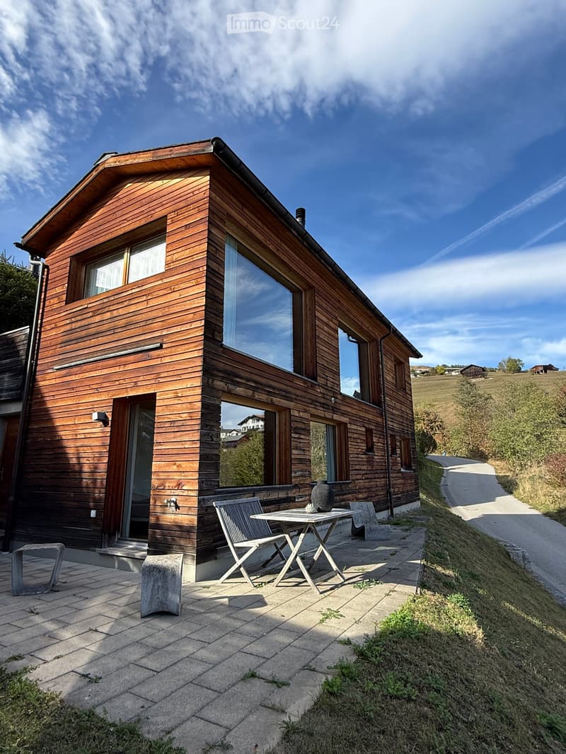 Modern wooden house with large windows and a raised ground floor, featuring a patio with a table and chairs.
