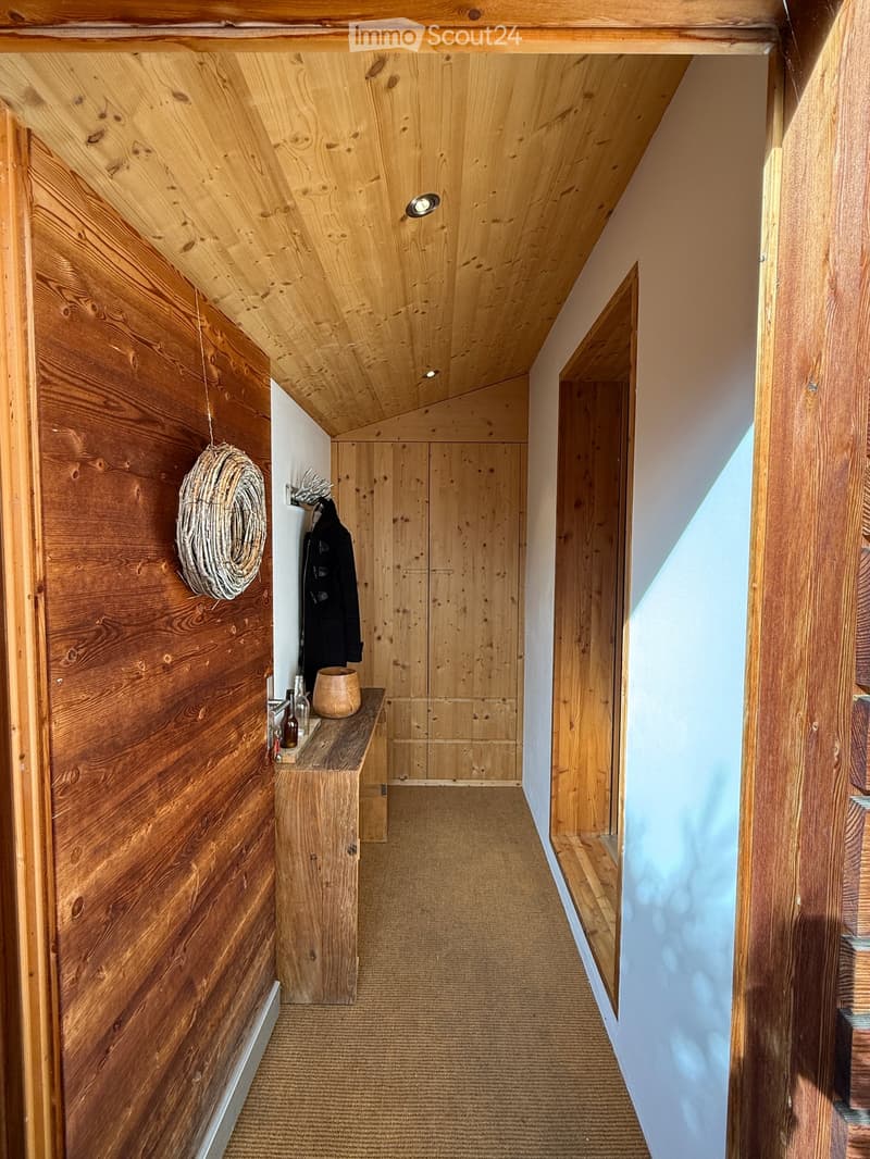 Wooden hallway with slanted ceiling, built-in wooden cabinetry, small wooden bench with decorative items, and coat rack with hanging coat.