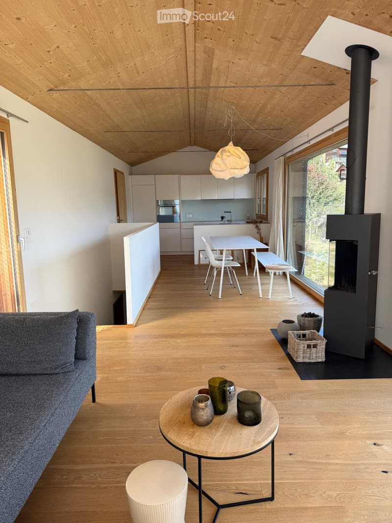 Open-plan living and dining area with wooden floors, a grey sofa, a round coffee table with decorative items, a white dining table with chairs, and a modern kitchen area with white cabinets and a stainless steel sink.