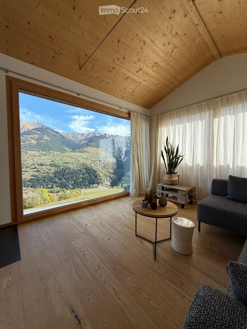 Modern living room with wooden floors, large panoramic window showcasing mountain view, sofa, coffee table, small round table, plant, and TV stand.