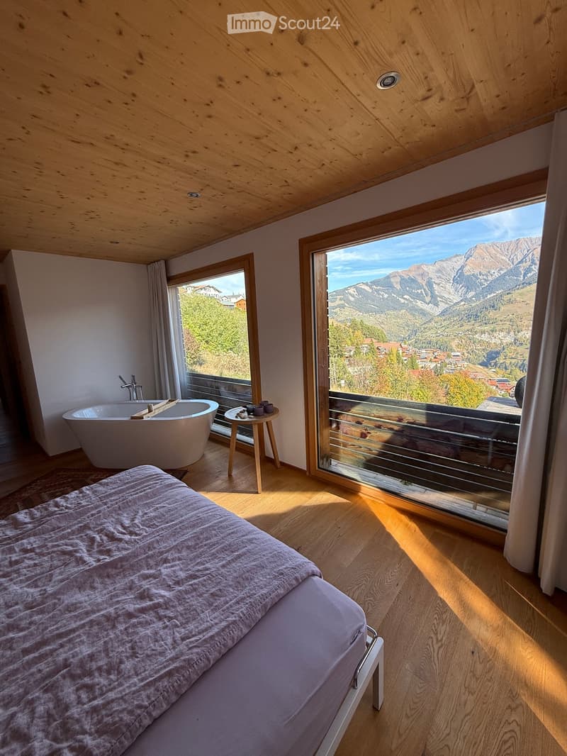 Bright bedroom with wooden floors, a large window offering a mountain view, a bed, a bathtub, and a small table with cups.