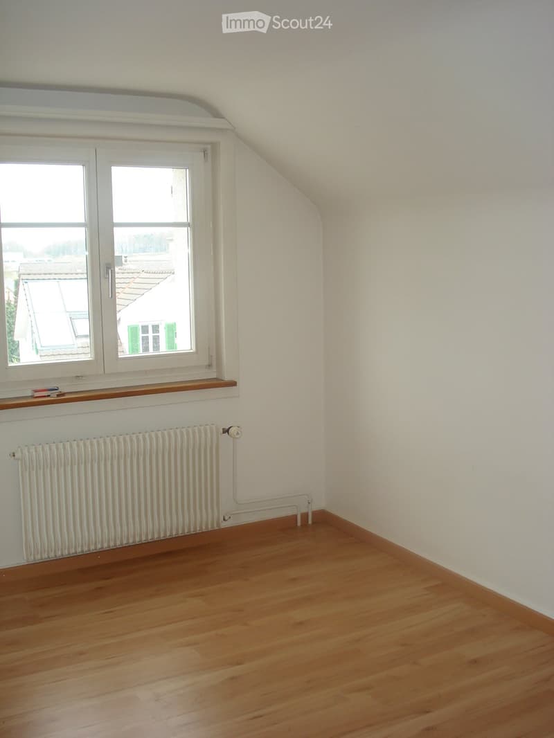 Empty attic room with wooden flooring, white walls, a radiator, and a window with a wooden shelf above it.