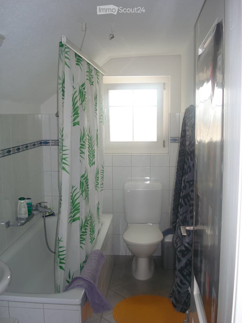 Bathroom with white tiles, a toilet, bathtub, sink, shower curtain with green leaf pattern, and a small orange stool.