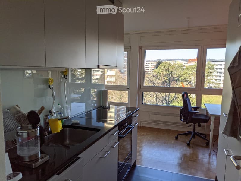 Modern kitchen with wooden flooring, large windows, sink, oven, coffee maker, and a small office area with a chair and table.