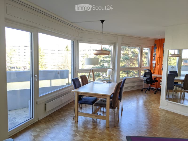 Bright room with large windows, wooden floor, a dining table with chairs, a lamp, and an office area with a desk and chair.