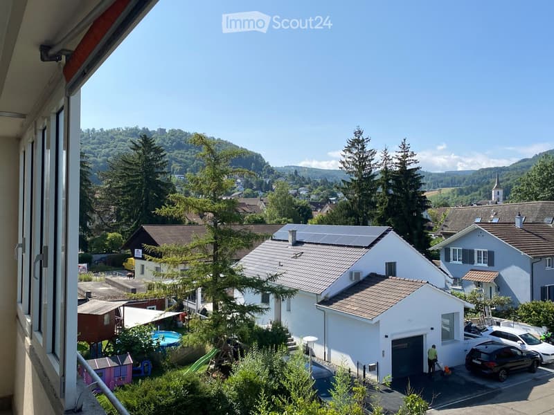 View from a balcony showing a residential area with houses, a swimming pool, a garage, parked cars, and a scenic view of hills and trees.