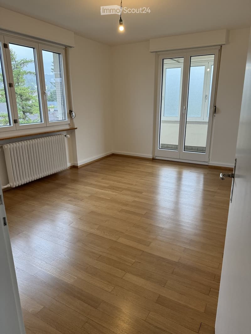 Empty room with light wooden flooring, white walls, large windows, and a radiator. No furniture or equipment present.