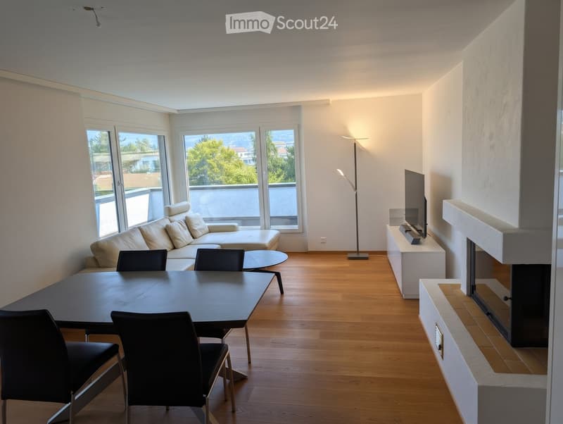 Modern living room with a white sofa, coffee table, dining table with chairs, television, floor lamp, and fireplace. Large windows offer a scenic view.